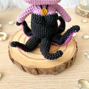 May include: A crocheted amigurumi doll of Ursula from The Little Mermaid. The doll is purple with black tentacles and a gold necklace. The doll is sitting on a wooden slice.