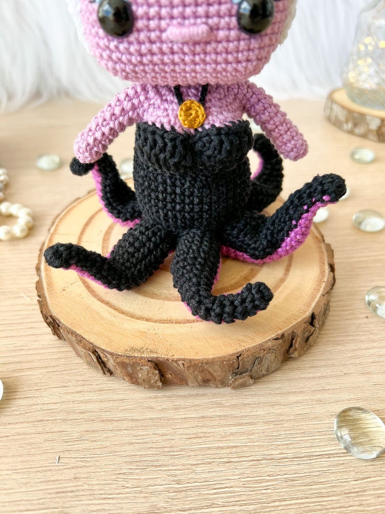 May include: A crocheted amigurumi doll of Ursula from The Little Mermaid. The doll is purple with black tentacles and a gold necklace. The doll is sitting on a wooden slice.