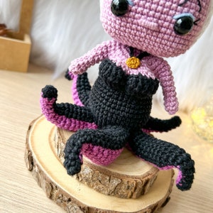 May include: A crocheted Ursula doll with purple skin, black hair, and a black and purple octopus tail. The doll is wearing a black and purple dress and a gold necklace. The doll is sitting on a wooden base.