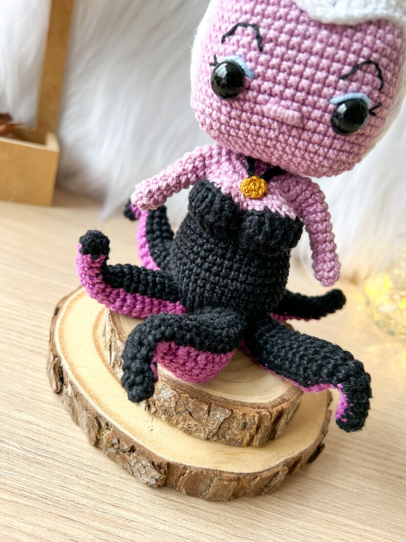 May include: A crocheted Ursula doll with purple skin, black hair, and a black and purple octopus tail. The doll is wearing a black and purple dress and a gold necklace. The doll is sitting on a wooden base.