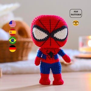 Może przedstawiać: Szydełkowa lalka Spider-Man w kolorach czerwonym, niebieskim i czarnym. Lalka ma maskę z białymi detalami oczu i kostium z emblematem pająka. Obraz zawiera małą grafikę z napisem "PDF PATTERN".