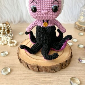 May include: A crocheted amigurumi doll of Ursula from The Little Mermaid. The doll is pink with a white hairpiece and black tentacles. The doll is wearing a black dress with a gold necklace.