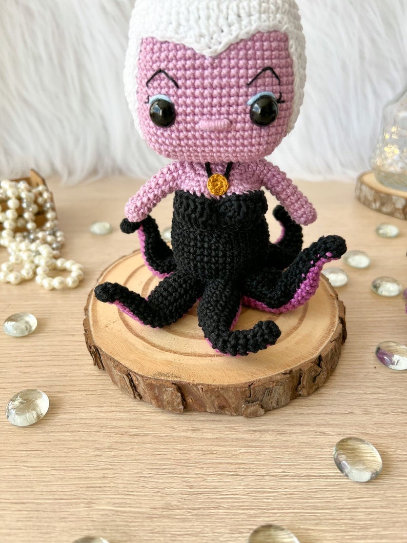 May include: A crocheted amigurumi doll of Ursula from The Little Mermaid. The doll is pink with a white hairpiece and black tentacles. The doll is wearing a black dress with a gold necklace.