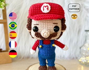 May include: Crocheted amigurumi toy of the video game character Mario. The toy is wearing a red hat with a white 'M' on it, a red shirt, blue overalls, and brown shoes. The toy has a brown mustache and a beige face.