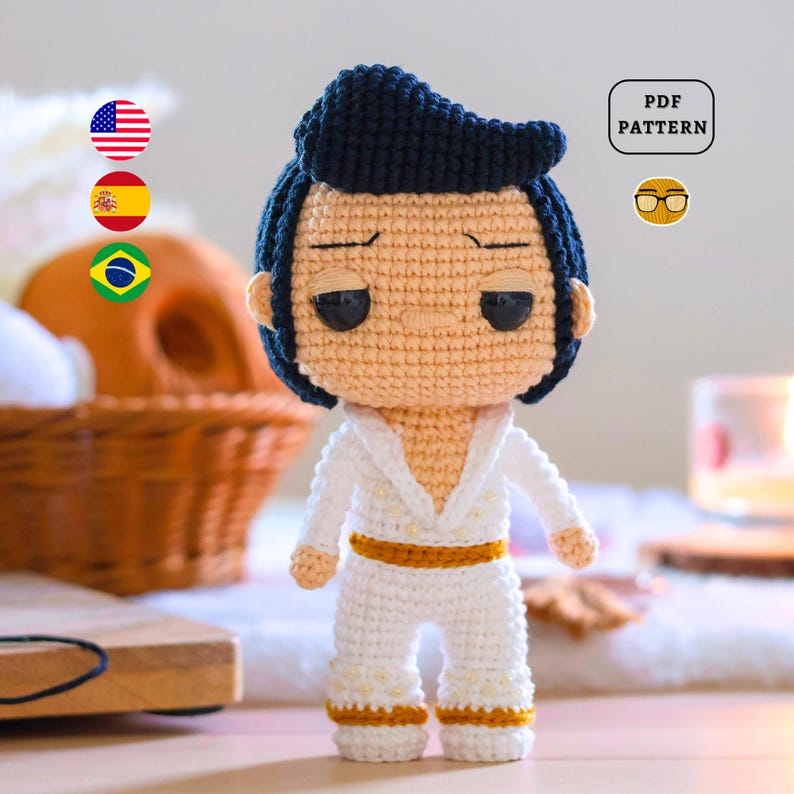 May include: Crocheted amigurumi doll of Elvis Presley in a white jumpsuit with a gold belt and black hair. The doll has a cartoonish style with large eyes and a serious expression. The doll is standing on a wooden surface.