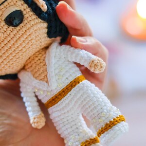 May include: A crocheted doll wearing a white jumpsuit with gold trim and white beads. The doll has black hair and brown eyes.