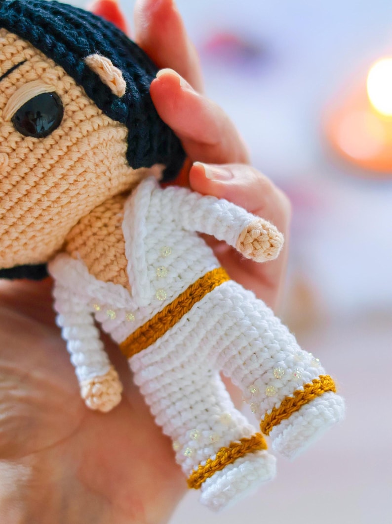May include: A crocheted doll wearing a white jumpsuit with gold trim and white beads. The doll has black hair and brown eyes.