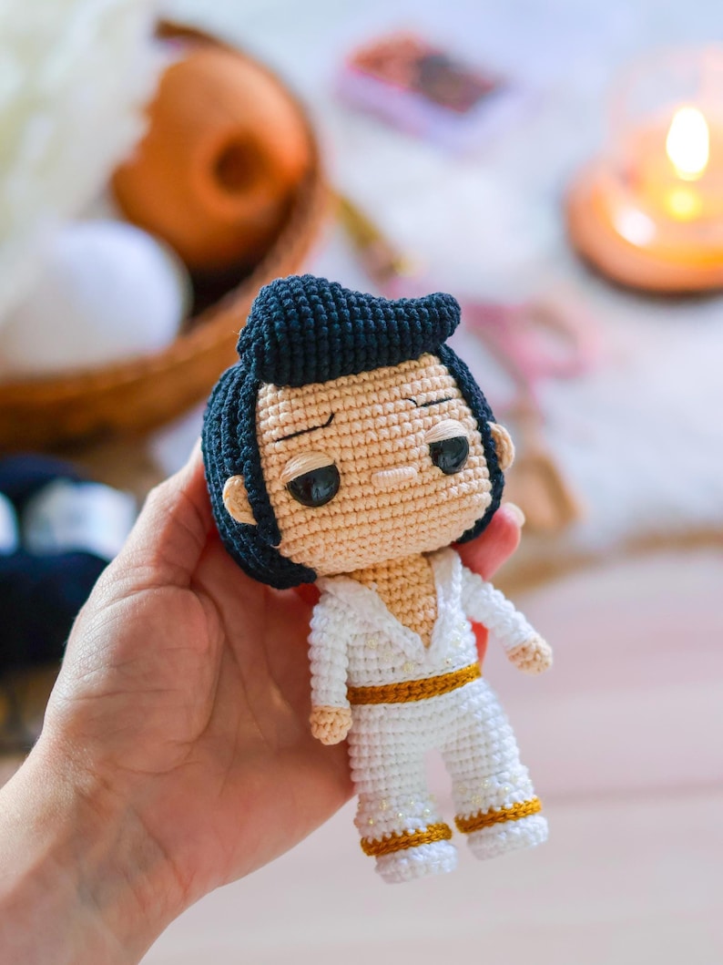 May include: A crocheted doll with black hair, a white outfit, and a gold belt. The doll has a serious expression on its face.