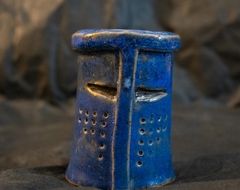 Puede incluir: Una escultura de cerámica con forma de yelmo de caballero, acabada con un esmalte azul brillante y profundo. La pieza presenta una abertura de visera en forma de T, ranuras oculares horizontales y orificios de ventilación circulares.
