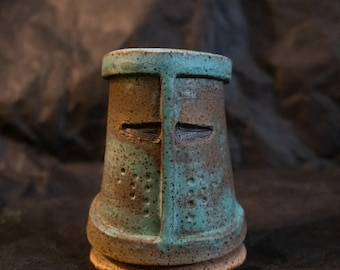 Puede incluir: Una taza de cerámica con forma de yelmo de caballero medieval. El recipiente presenta un esmalte moteado en tonos verde azulado y marrón, con una abertura en forma de T para la visera, un borde superior plano y una base ensanchada.