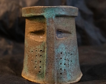 Puede incluir: Una taza de cerámica con forma de yelmo de caballero medieval. El recipiente presenta un esmalte moteado en tonos marrón y verde azulado, una abertura de visera en forma de T y dos secciones verticales en la parte inferior con perforaciones.