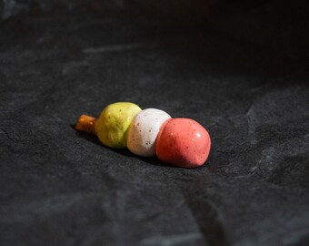 Puede incluir: Una brocheta dango de cerámica con tres esferas redondeadas y moteadas en verde lima, blanco y rosa coral. Las esferas están dispuestas en una fila lineal, con una pequeña parte de un palillo de madera marrón visible en la esfera verde.