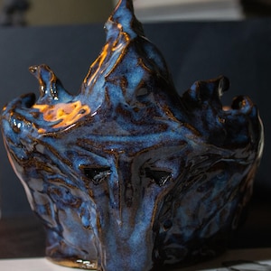 Peut inclure: Sculpture en céramique artisanale au design abstrait unique. La pièce présente un glaçage bleu profond avec des accents bruns, créant un aspect texturé et organique. La sculpture a une forme distinctive, avec un sommet pointu et des ouvertures ressemblant à des yeux.
