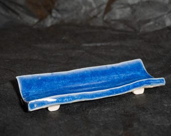 Puede incluir: Una bandeja rectangular de cerámica con un acabado esmaltado en azul vibrante y una superficie ligeramente curva y cóncava. La bandeja presenta un aspecto brillante y texturizado, apoyada sobre pequeñas patas blancas en forma de bloque.