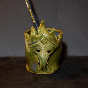 Op de afbeelding: Een handgemaakte keramische potloodhouder in groen- en blauwtinten. De houder heeft een gestileerd gezichtsontwerp met puntige kenmerken en zwarte ooguitsparingen. Een zwart potlood met witte tekst staat erin.