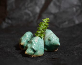 Puede incluir: Tres pequeñas figuras de cerámica esculpidas a mano con un esmalte turquesa claro y sutiles tonos marrones. Cada figura presenta un hocico puntiagudo y orejas redondeadas, parecidas a un animal pequeño. Una rama de helecho verde se asoma detrás.