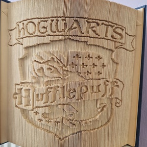 May include: A book folded into a 3D sculpture of the Hogwarts crest. The crest features a lion, eagle, badger, and snake, with the words "Hogwarts" and "Hufflepuff" in a banner.