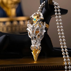May include: A silver and gold pendant on a silver ball chain. The pendant features an intricate, openwork silver frame with swirling patterns, a central gold-toned element, and a small, round, bright turquoise stone set near the top bail.
