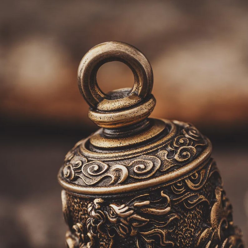 May include: Close-up of a bronze-colored bell with intricate dragon and cloud designs. The bell has a loop at the top for hanging and detailed carvings around the body. The metal has a slightly aged appearance.