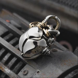 May include: A silver pendant with a futuristic, mechanical design. The pendant has a rounded, segmented body with gold-colored accents and a loop for a chain. The overall aesthetic is reminiscent of science fiction or steampunk.