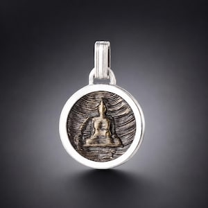 Puede incluir: Un colgante circular que presenta una figura central de tono dorado en una pose de meditación sentada. El fondo dentro del círculo tiene un patrón oscuro, texturizado y ondulado. El colgante está enmarcado en plata pulida e incluye una reasa vertical.