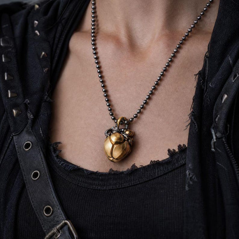 May include: A metallic pendant shaped like a scarab beetle with a polished gold-tone finish, suspended from a dark gunmetal-colored ball chain necklace. The beetle features intricate dark metal detailing around the top and a smooth, rounded body.