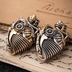 May include: Two heart-shaped pendants crafted from metallic material, featuring intricate mechanical details like gears, pipes, and vents. The design combines gold and silver tones, showcasing a complex, layered structure with a prominent circular fan element.
