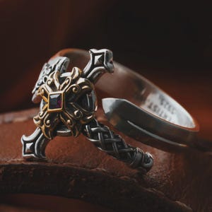 Puede incluir: Anillo de plata con un diseño de cruz decorativa. La cruz presenta un centro dorado con una gema cuadrada de color rojo oscuro. El anillo tiene detalles intrincados y una caña dividida.