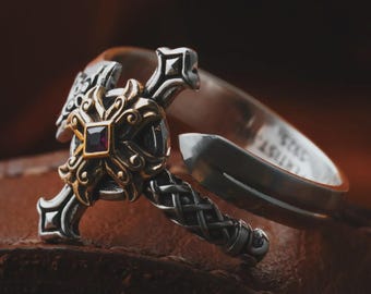 May include: A silver ring with a decorative cross design. The cross features a gold-colored center with a square, dark red gemstone. The ring has intricate detailing and a split shank.