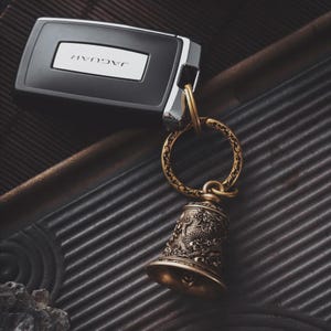 May include: A Jaguar car key with a decorative brass bell keychain. The bell features an ornate design and is attached to a circular ring. The key is black with a silver accent and the Jaguar logo.