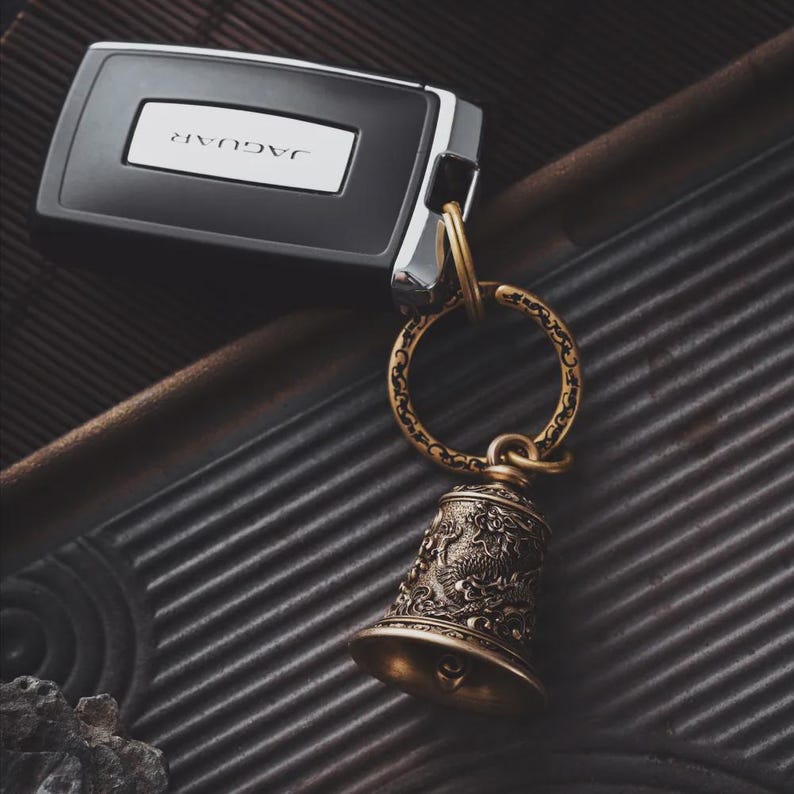 May include: A Jaguar car key with a decorative brass bell keychain. The bell features an ornate design and is attached to a circular ring. The key is black with a silver accent and the Jaguar logo.