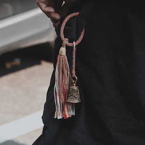 May include: A decorative bell charm with a tassel and braided loop. The tassel features red, orange, white, and blue threads. The bell is bronze-colored with intricate patterns. The charm is attached to a black strap.