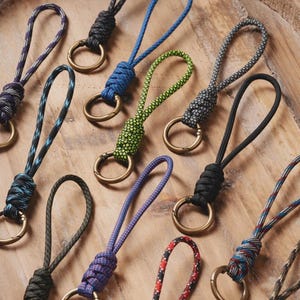 Può includere: Una collezione di portachiavi in paracord colorati con anelli in metallo color ottone. I portachiavi presentano vari motivi e colori, tra cui blu, verde, nero e viola. I portachiavi sono disposti su una superficie di legno.