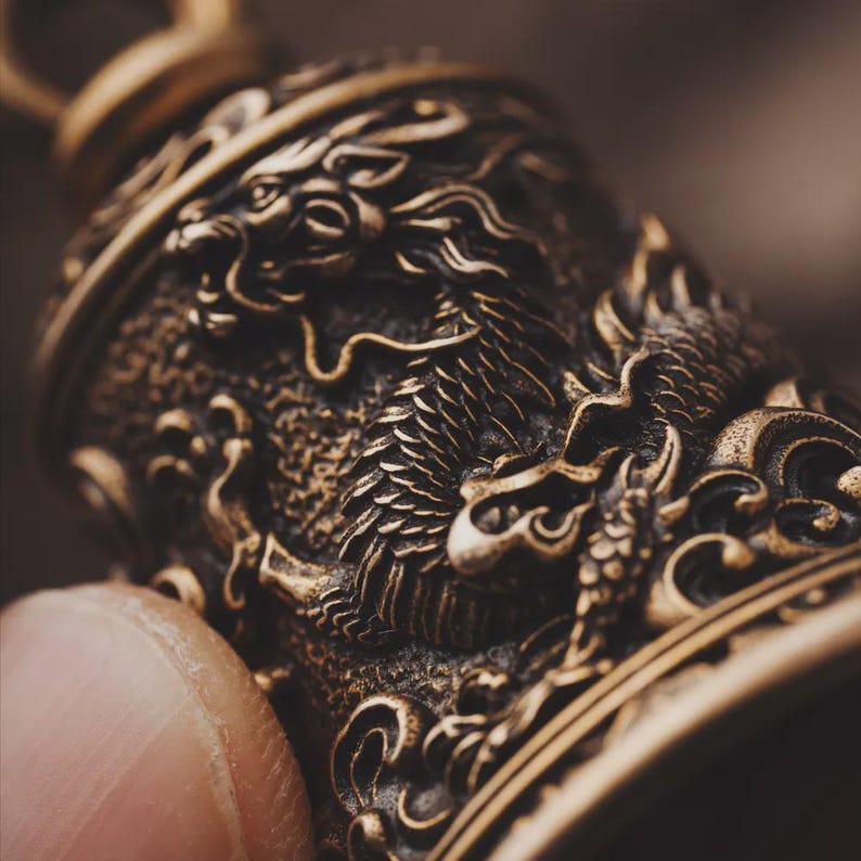 May include: Close-up of a bronze-colored bell with an intricately carved dragon design. The bell features detailed relief work, showcasing the dragon's scales, mane, and facial features. The bell is suspended from a loop at the top.