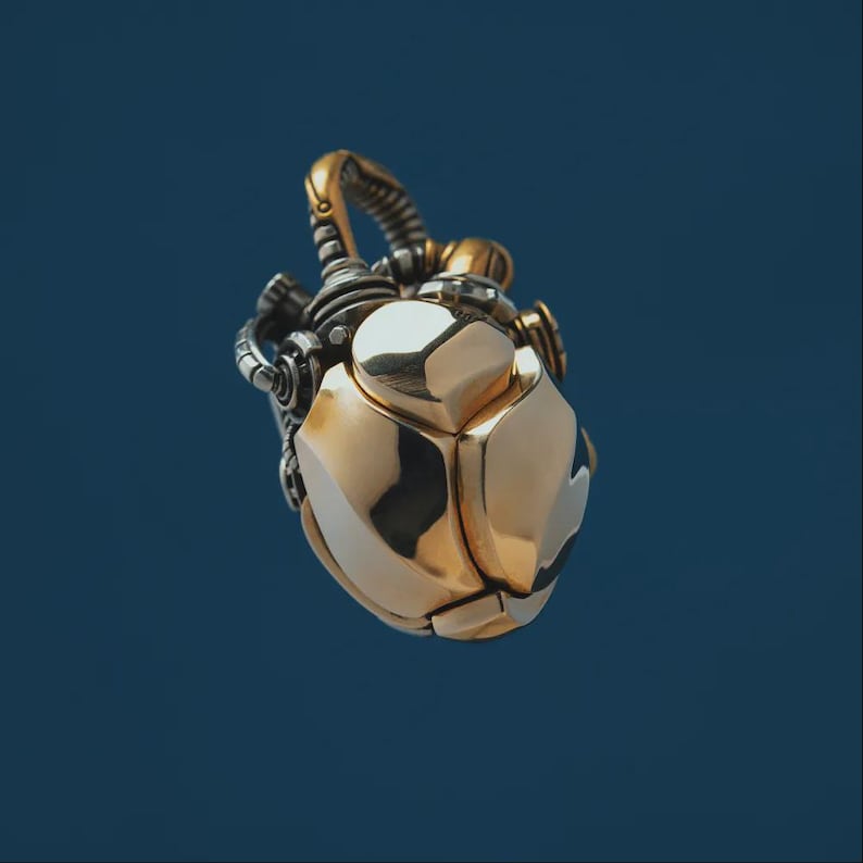 May include: A metallic pendant shaped like a mechanical heart, featuring a polished gold-tone finish on the main body and intricate silver-tone gear-like details at the top. The design incorporates smooth, rounded plates and visible structural joints.