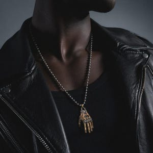 May include: A silver-toned ball chain necklace featuring a pendant shaped like a mechanical hand. The pendant has a metallic finish with segmented finger details and a gold-colored accent at the wrist joint, suspended from a simple metal clasp.