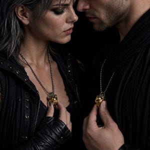 May include: A gold-toned heart-shaped pendant with intricate metallic detailing, suspended from a silver-colored ball chain necklace. The pendant features a smooth, polished finish and a structured design, worn as a matching set by two individuals.