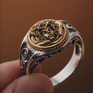May include: A silver ring with a gold-colored medallion featuring a detailed face design. The ring has intricate scrollwork on the sides and a small, dark-colored stone set in the center of the medallion. The ring is held by a person's finger.