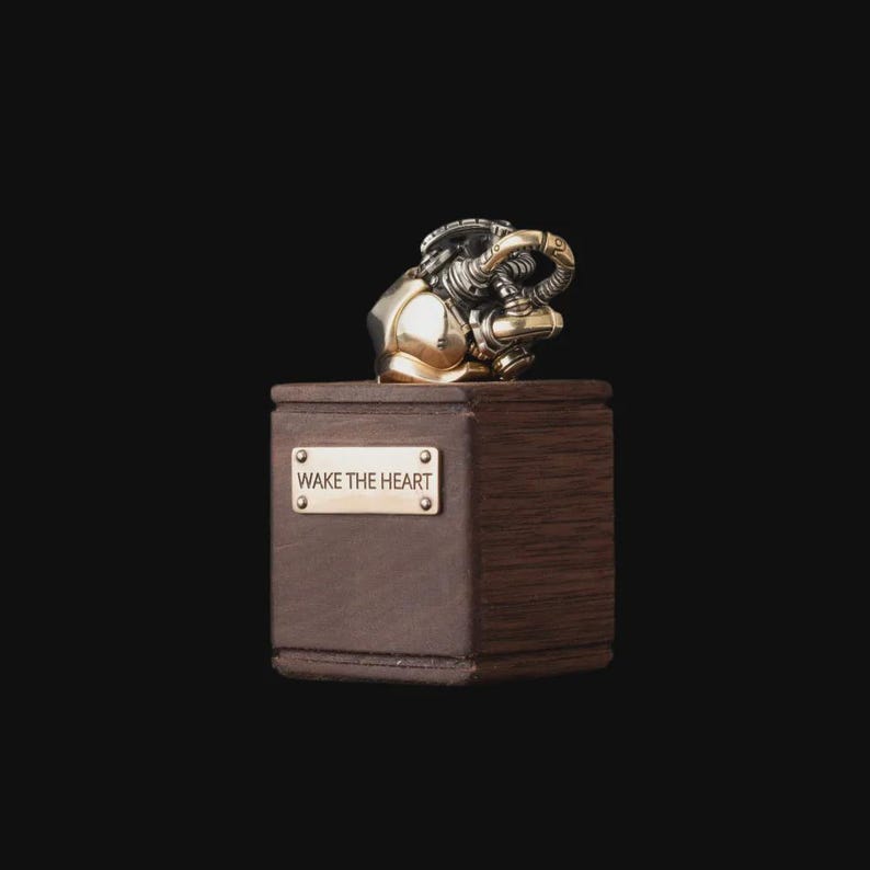 May include: A metallic anatomical heart sculpture with gold and silver tones resting on a dark brown wooden block. A rectangular brass plaque is attached to the front of the block, featuring the engraved text WAKE THE HEART in capital letters.