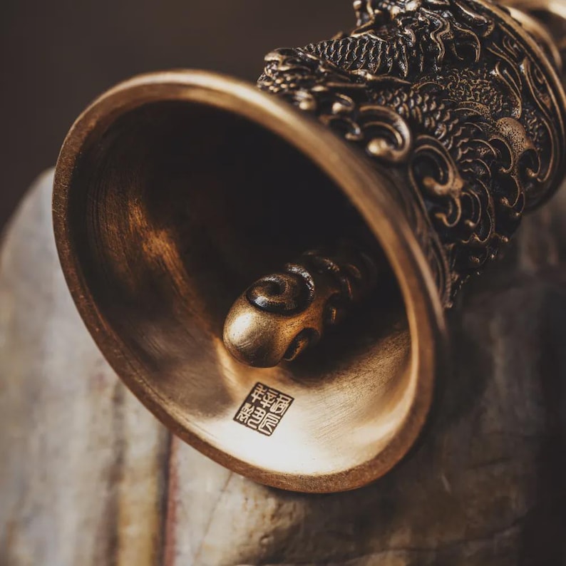 May include: Close-up of a bronze bell with intricate dragon detailing on the handle. The bell has a smooth, polished finish and a small, decorative clapper inside. A square stamp is visible near the base of the bell.