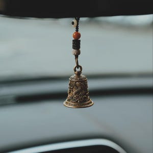 May include: A bronze-colored bell with intricate dragon and floral designs, suspended by a chain and a string of beads. The bell is likely a decorative item, possibly for a car or home decor.