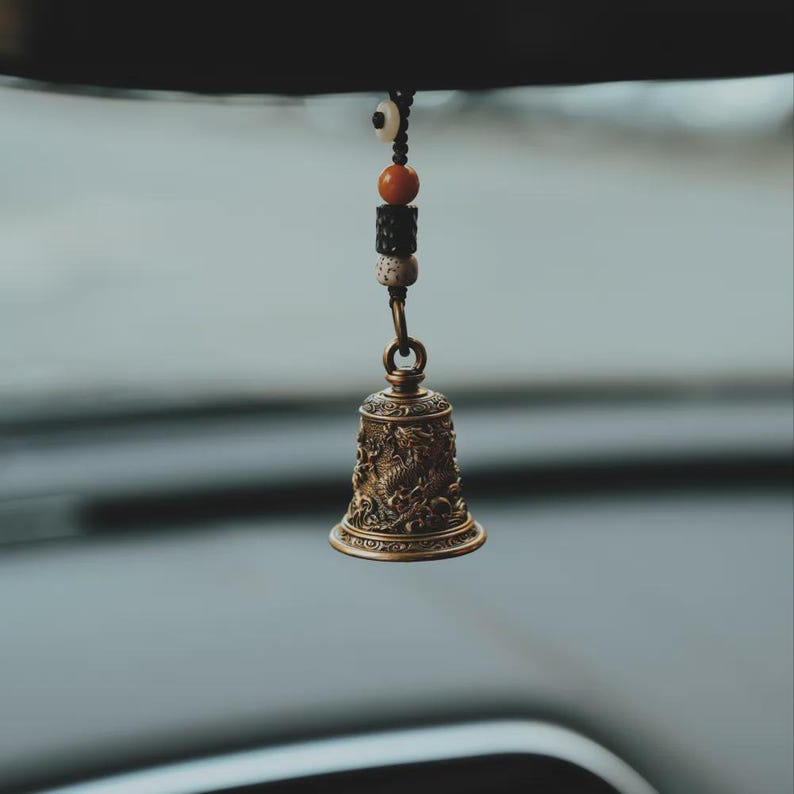 May include: A bronze-colored bell with intricate dragon and floral designs, suspended by a chain and a string of beads. The bell is likely a decorative item, possibly for a car or home decor.