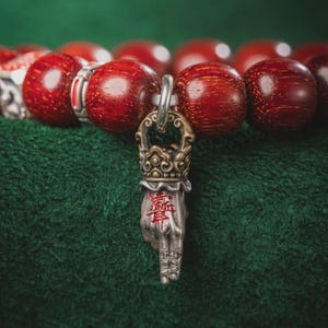May include: A close-up of a wooden bead bracelet with a silver charm. The beads are a deep red color. The charm is a hand with a crown and Chinese characters. The background is a green, textured fabric.