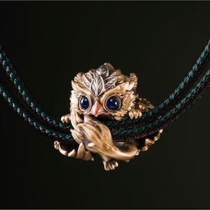 Könnte beinhalten: Eine detaillierte, goldene Eulenfigur mit blauen Augen und einem roten Schnabel, die auf einer dunkelgrünen und braunen geflochtenen Schnur sitzt. Die Eule ist aufwendig mit blattartigen Details an Kopf und Flügeln gestaltet. Der Hintergrund ist dunkel.