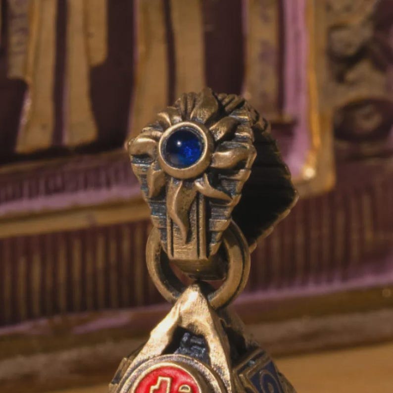 May include: Close-up of a decorative, ornate pendant. The pendant features a gold-colored metal design with a blue gemstone at the top. The design includes floral and geometric elements, with a circular ring and a detailed lower section.