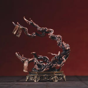 May include: A bronze-colored, stylized tree sculpture with intricate details. The sculpture features a twisted trunk and branches, with small rectangular elements hanging from the branches. It sits on a square base with ornate carvings, set against a deep red background.
