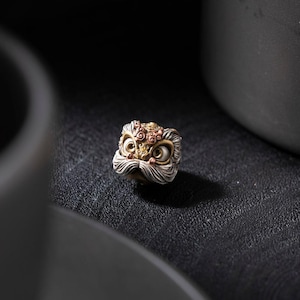 May include: A silver lion dance head charm with gold and copper accents. The lion's face is detailed with intricate carvings and has large, round eyes.