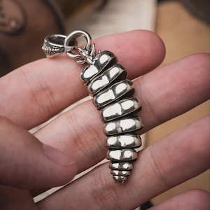 May include: A polished silver pendant shaped like a segmented rattlesnake tail. The piece features a series of curved, stacked metallic segments that taper towards the bottom, topped with a sturdy integrated bail for attachment to a chain or cord.