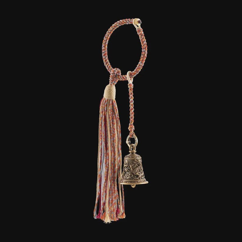 May include: A decorative bronze bell with intricate carvings hangs from a braided cord and tassel. The tassel features a mix of red, orange, and blue threads. The bell is suspended from a loop, creating a unique and ornate design.