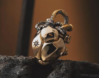 May include: A gold-colored pendant in the shape of a heart, with intricate mechanical details and a star-shaped design. The pendant has a silver-colored bail and is set on a dark, textured surface.